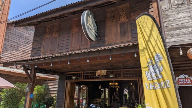 Wooden restaurant exterior with a circular sign, yellow Singha flag, and Stella Artois sign, plants outside, and open door.