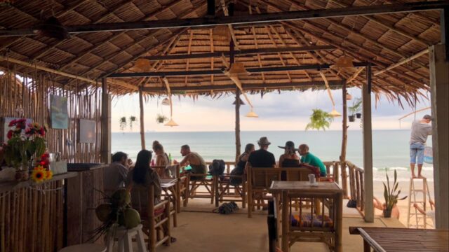 People sit at wooden tables in a thatched beachside café, facing the ocean with a relaxed, tropical atmosphere.