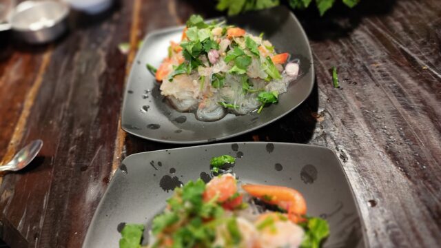 Two black plates with a fresh salad of shrimp, herbs, and vegetables on a wooden table, with greens in the background.