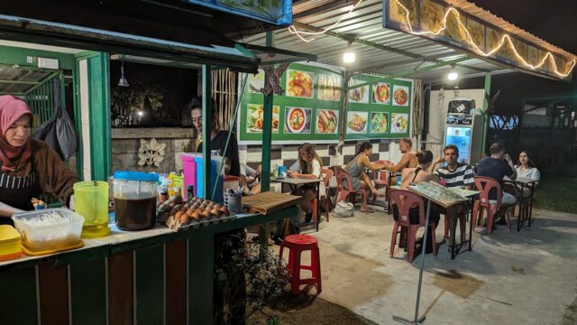 Outdoor night food stall with people dining at tables, a woman preparing food, and menus displayed on the wall.