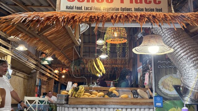Colorful food stall named Sunee Place sells fruit, Nutella, and drinks with tropical decor under a thatched roof.