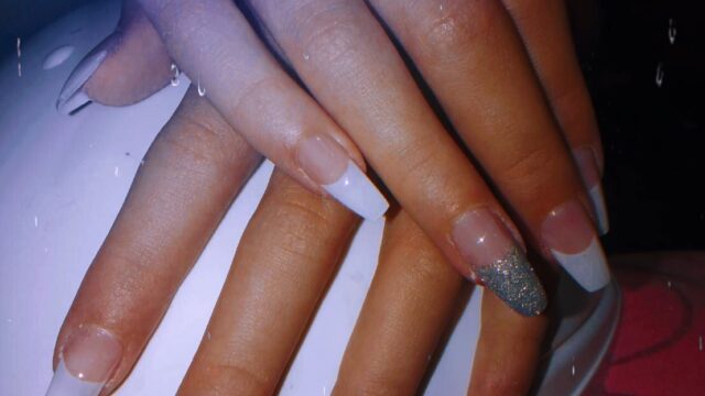 Hands with long, square French tip nails, two with silver glitter, resting on a white object with a pink background.