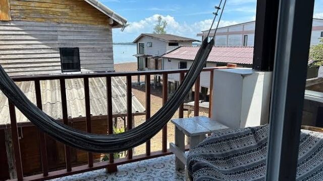A hammock on a balcony overlooks wooden buildings and the sea under a bright blue sky with clouds.