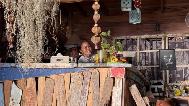 A smiling person stands behind a rustic wooden bar lined with wine bottles, with plants and hanging decor in a cozy, eclectic space.