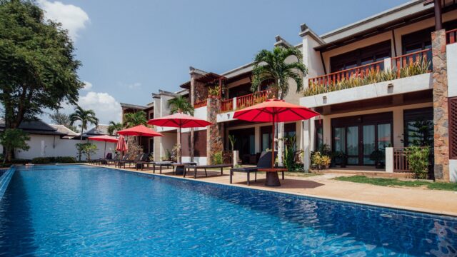 Large pool beside a modern villa with red umbrellas, lounge chairs, palm trees, and a clear blue sky.
