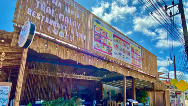 A bamboo Thai-Malay restaurant with a large menu sign, blue sky, and outdoor seating under a canopy.