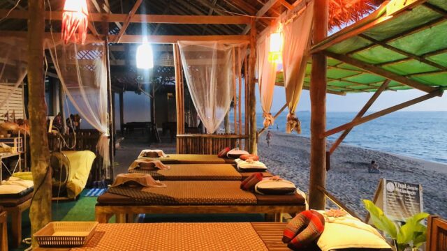 Open-air beachside spa with massage beds, pillows, and soft lighting, overlooking sand and ocean at sunset.