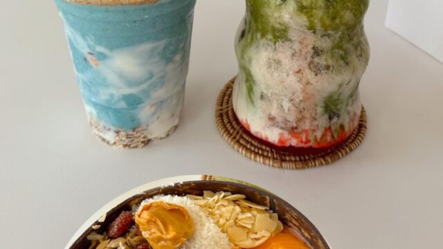 A vibrant smoothie bowl with fruit and seeds, a blue smoothie, and a green iced drink on a white table.