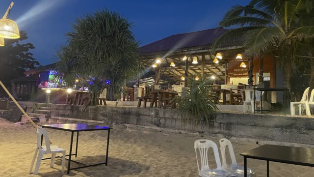Outdoor beach restaurant at dusk with empty tables, plastic chairs on sand, and warm lights glowing under a wooden roof.