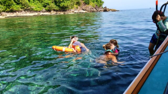 Three people snorkeling in clear water near a lush, green shoreline, with one person holding an orange float.