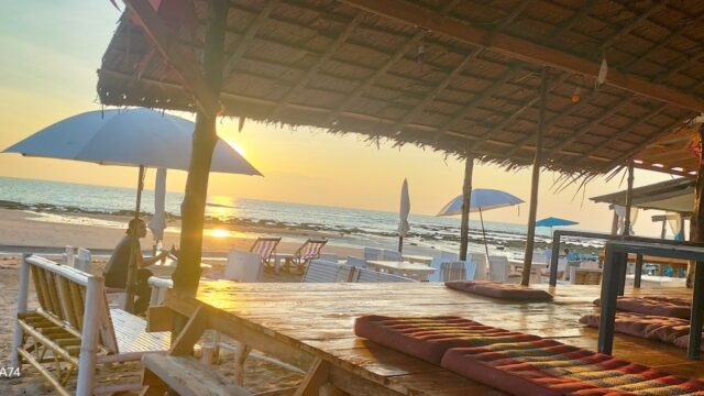 Sunset view from a beachside hut with mats, wooden seating, umbrellas, and a person enjoying the scenery by the shore.