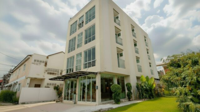 A modern four-story building with large windows, surrounded by greenery, under a partly cloudy sky.