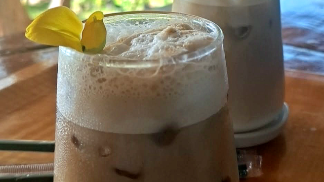 Two iced coffee drinks on a wooden table, each garnished with a yellow flower, with a green outdoor background.