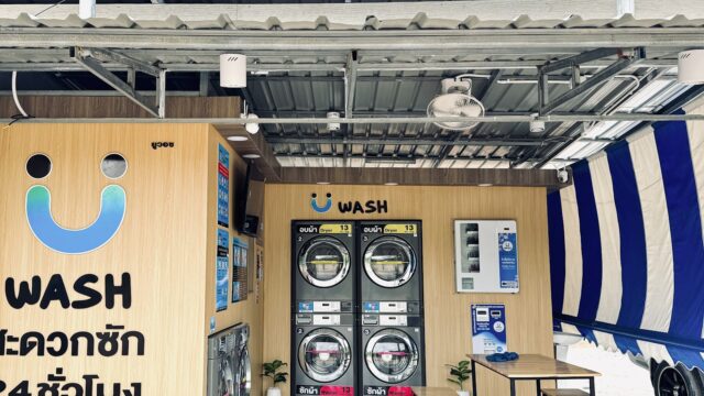A small self-service laundromat with washing machines, vending machines, benches, and blue-and-white awnings. Signs are in Thai and English.