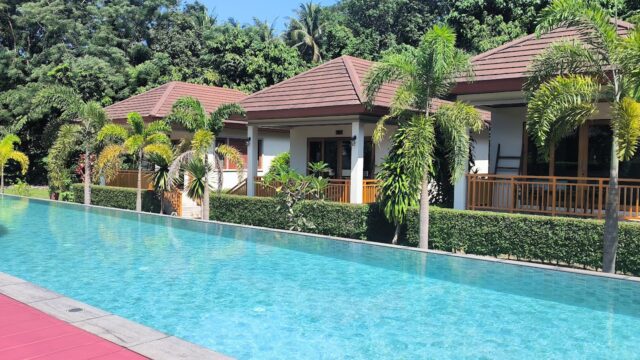 Long swimming pool in front of modern villas with tropical plants and palm trees under bright sunlight.