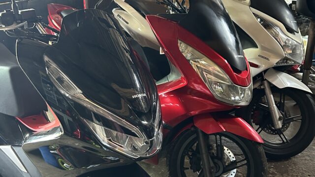 Three Honda scooters—black, red, and white—are parked indoors near tires and boxes, with a mesh fence in the background.