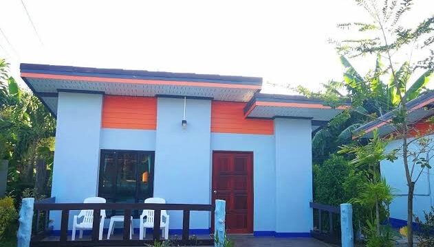 Small modern house with an orange roof, porch with white chairs, and lush greenery surrounding the yard.